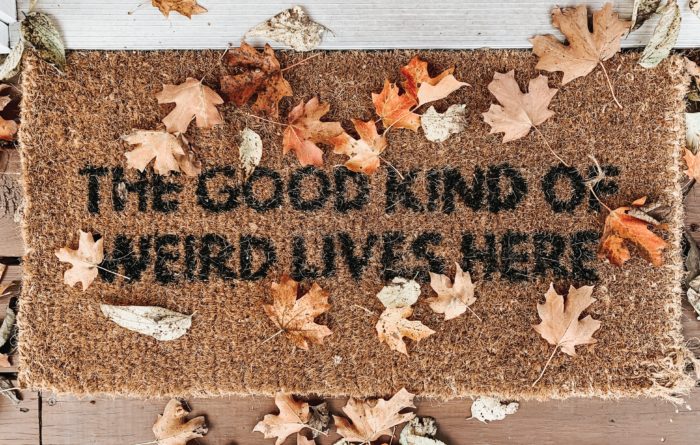 front door mat with fall leaves scattered around it, with text that says the good kind of weird lives here