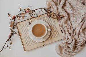 open book with a cup of tea in the center next to a beige sweater and a sprig of fall flowers